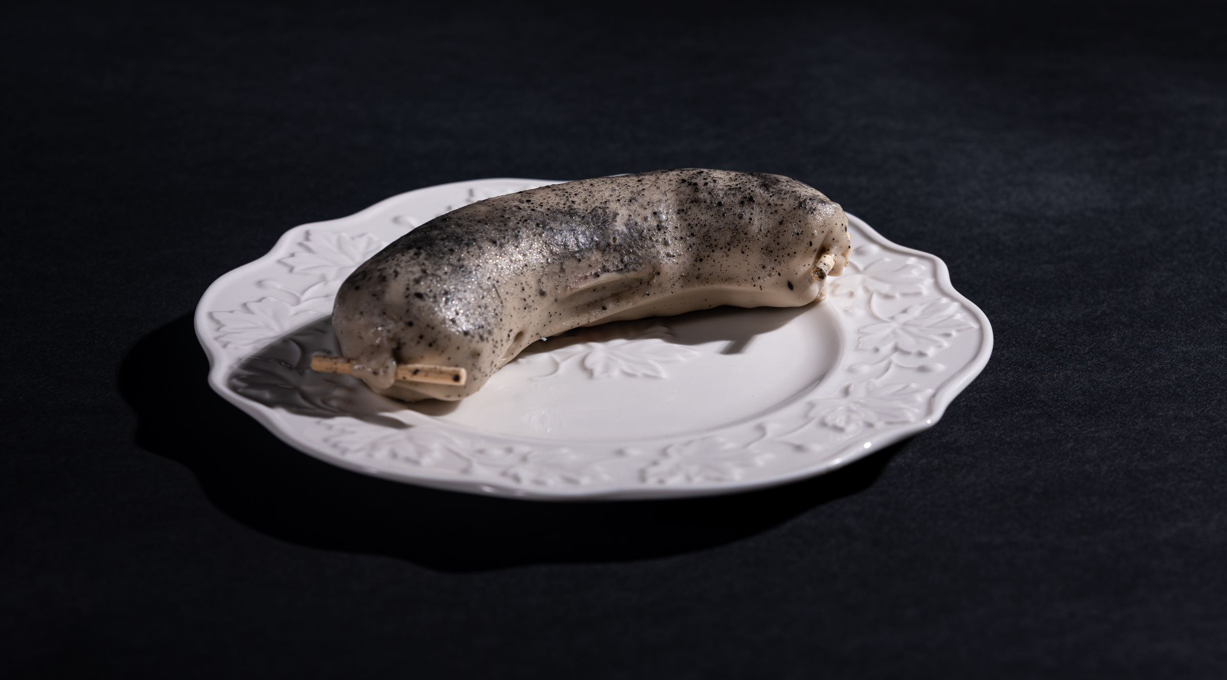 Tečkovaná jitrnice na reliéfním porcelánovém talíři vystupuje ze tmy — dezert v přestrojení